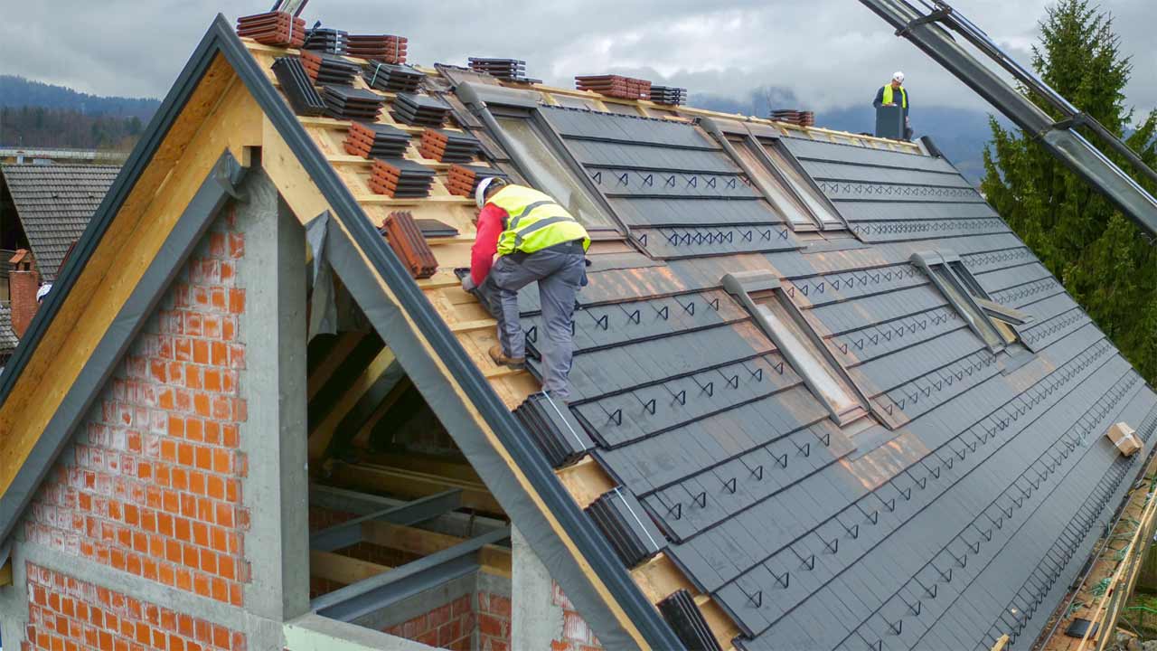 Workers in safety vests are installing roof tiles on a sloped roof of a house under construction, likely factoring in the Value Calculator for optimal efficiency. One worker is on the roof, while another stands at the edge. A mountainous landscape is in the background.